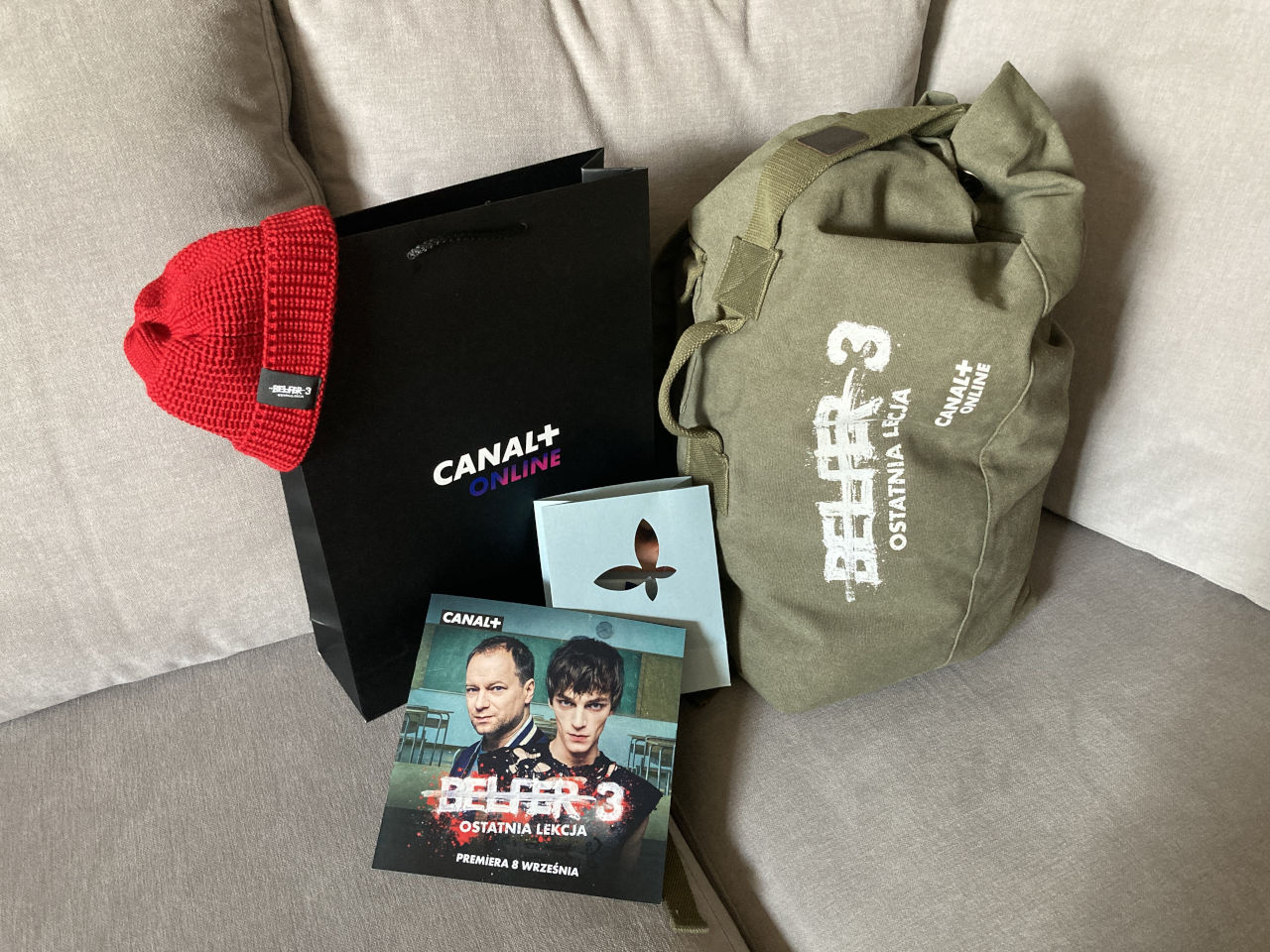 Zestaw prezentowy Canal+ Online przygotowany przez Deuter DLM – duża czarna torba papierowa, czerwona czapka, ulotka Belfer 3 Ostatnia Lekcja oraz zielony plecak podróżny z nadrukiem filmowym, zapakowane z konfekcją